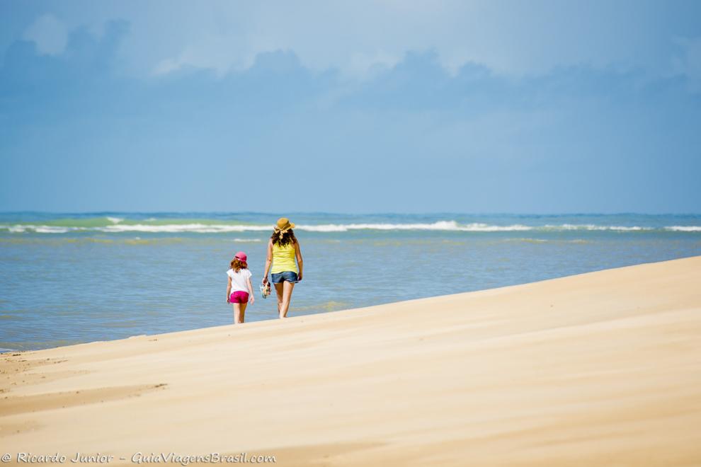 Imagem de duas pessoas andando nas areias claras da praia e ao fundo um mar maravilhoso.